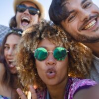 Group of friends having fun taking a selfie. They are all looking at the camera and smiling and laughing. Some are making funny faces. Background is blue sky on a sunny day.